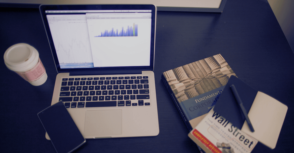 A laptop with stock market screen in a table along with some books about basic finance concepts like wall street and money in business